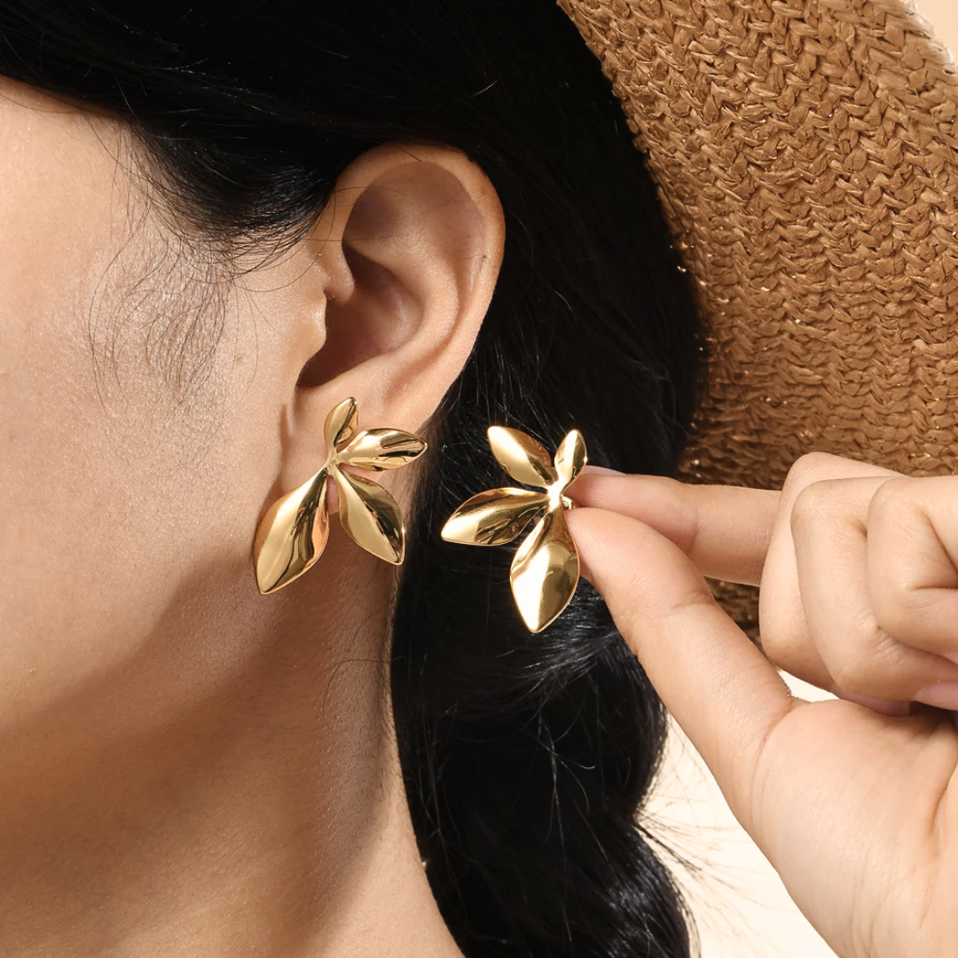Donna con capelli neri e cappello di paglia che indossa orecchini a lobo a fiore botanico in acciaio inossidabile placcato oro 18K, design scultoreo a petali multipli lucidi, vista laterale ravvicinata sul lobo — tendenza gioielli Primavera-Estate 2026