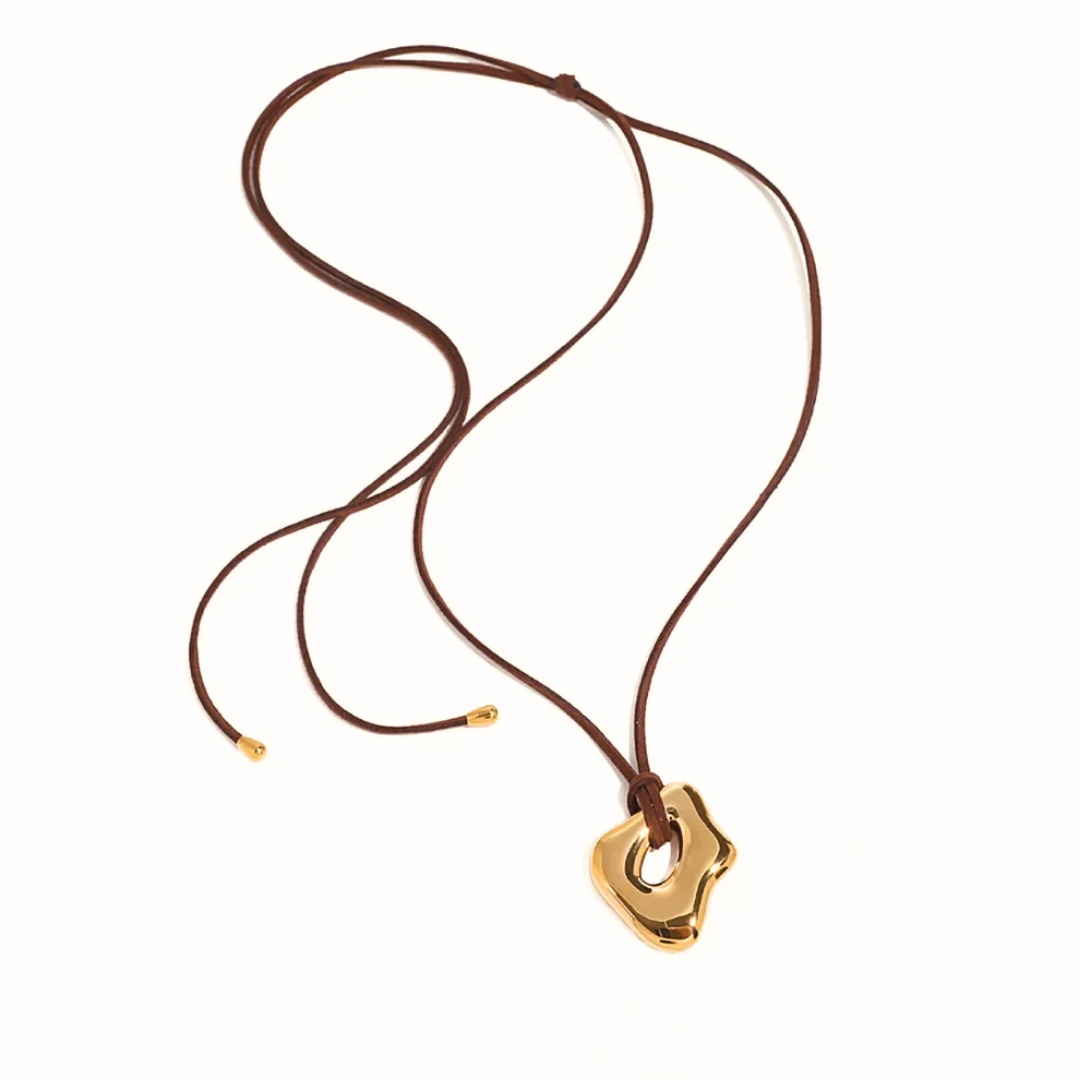 Collana con pendente dorato di forma organica irregolare su cordino marrone scuro.