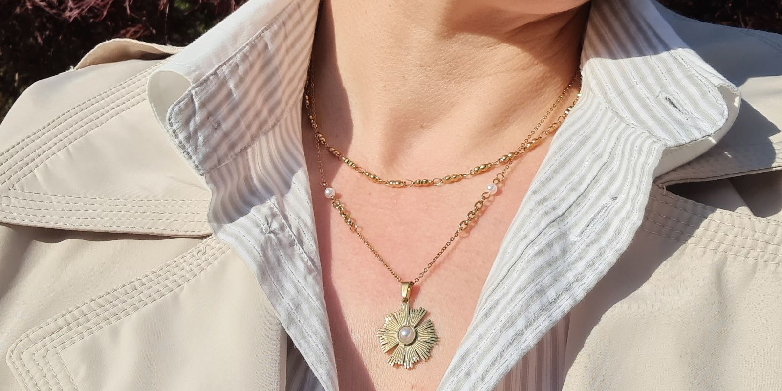 Primo piano di doppia collana in oro da donna indossata con outfit elegante. Girocollo sottile e collana con ciondolo a sole dorato e perla centrale.